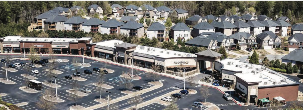 Aerial view of a shopping center with several stores and a partially filled parking lot, surrounded by residential houses and trees.